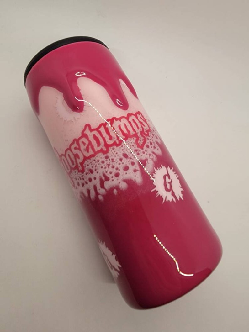 May include: A pink and white tumbler with a black lid. The tumbler has a white design with the words "Goosebumps" and a "G" with a white splatter effect.