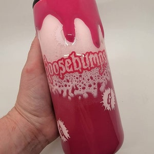 May include: A pink and white tumbler with a black lid. The tumbler has a white design of the words "Goosebumps" and a white "G" with a white splatter design.