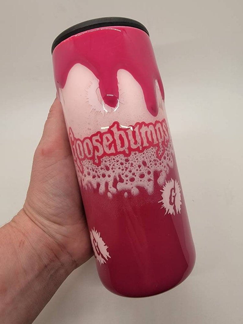 May include: A pink and white tumbler with a black lid. The tumbler has a white design of the words "Goosebumps" and a white "G" with a white splatter design.