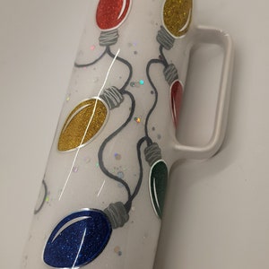 May include: White tumbler with a handle, featuring a Christmas lights design. The lights are red, yellow, blue, and green, with silver glitter accents.