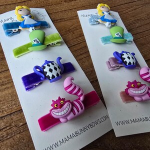 Op de afbeelding: Haarclips met een Alice in Wonderland-thema. De clips tonen personages als Alice, de hoed van de Mad Hatter, een theepot en de Cheshire Cat. Elke clip is bevestigd aan een fluwelen lint in verschillende kleuren. De website-adres is zichtbaar.