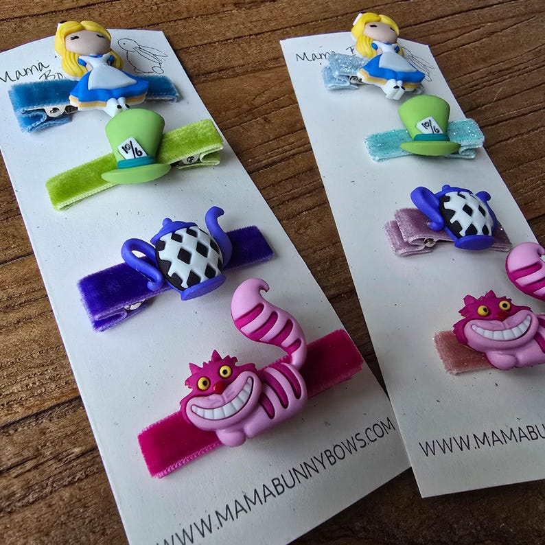 Op de afbeelding: Haarclips met een Alice in Wonderland-thema. De clips tonen personages als Alice, de hoed van de Mad Hatter, een theepot en de Cheshire Cat. Elke clip is bevestigd aan een fluwelen lint in verschillende kleuren. De website-adres is zichtbaar.