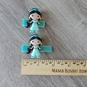 May include: Two turquoise blue hair clips with a princess character on each. The princess is wearing a turquoise blue outfit and has black hair. The clips are 1 inch long.