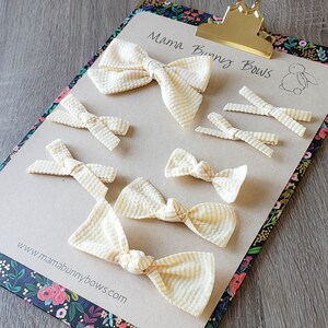 May include: A collection of yellow and white striped hair bows on a brown background. The bows are arranged in different sizes and styles. The text "Mama Bunny Bows" and "www.mamabunnybows.com" is visible on the background.