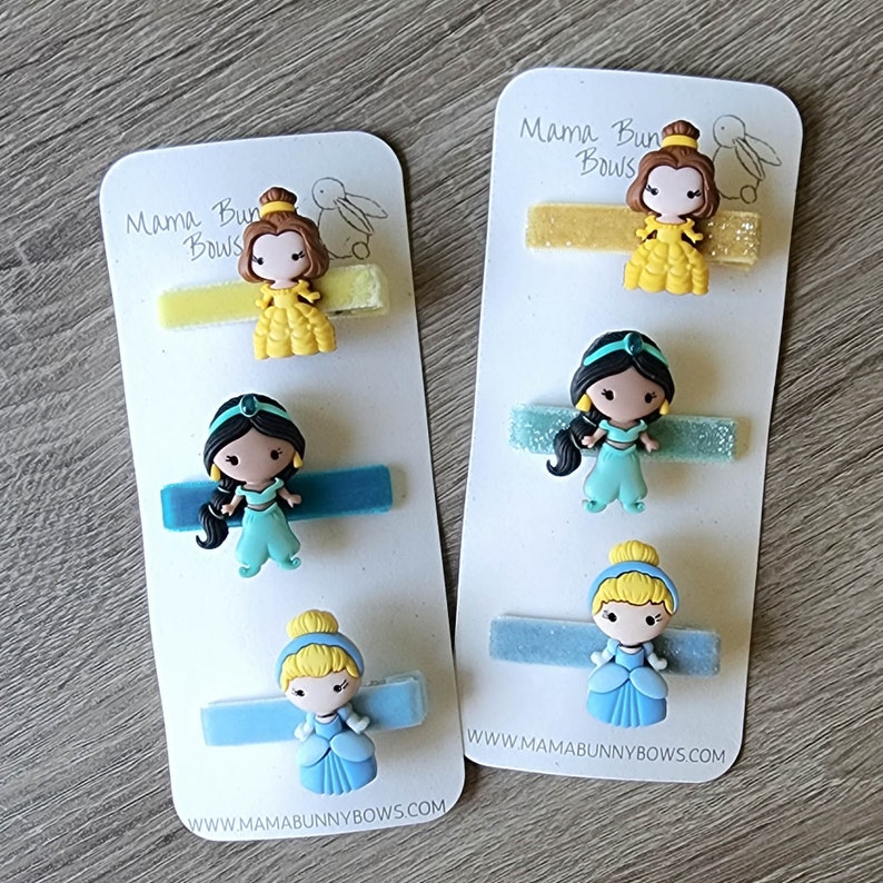 May include: Three sets of princess-themed hair clips on white cards. Each set includes a princess character and a glittery ribbon. The princesses are Belle, Jasmine, and Cinderella. The cards have the text "Mama Bun Bows" and a website address.