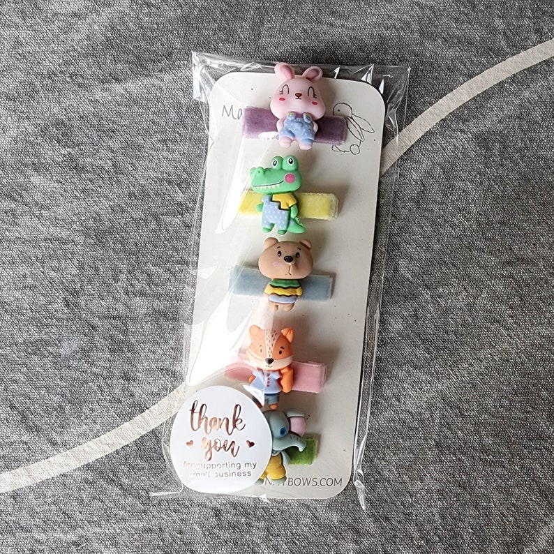 May include: A set of six colourful hair clips featuring various animal designs, including a bunny, crocodile, bear, fox, and elephant. The clips are packaged on a white card with the text "Thank you for supporting my small business" and the website address "BOWS.COM".