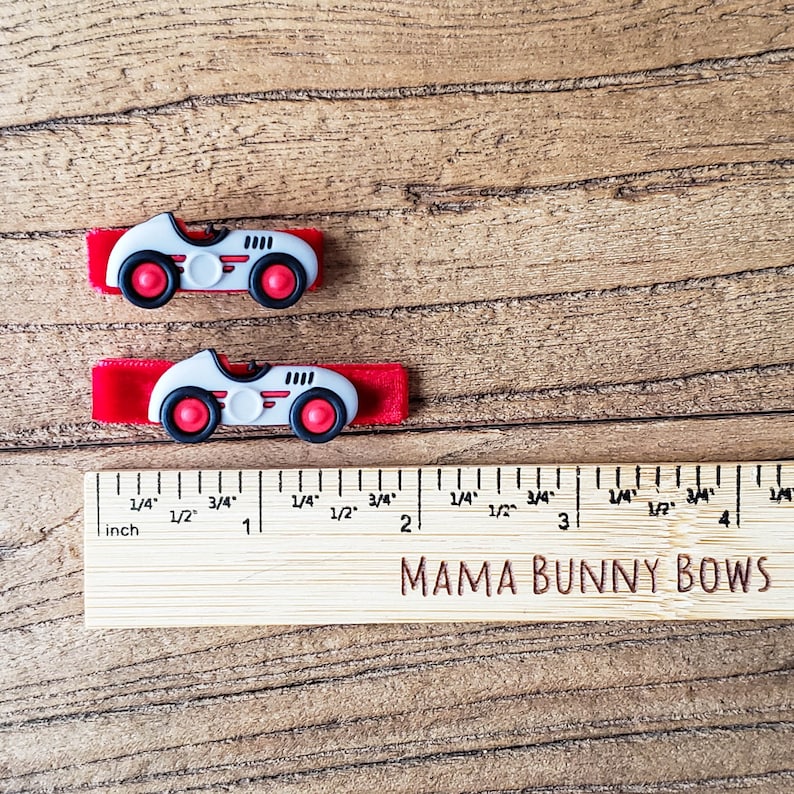 May include: Two white toy race cars with red wheels and red velvet ribbon attached. The cars are about 1 inch long. A wooden ruler with the text "MAMA BUNNY BOWS" is below the cars.