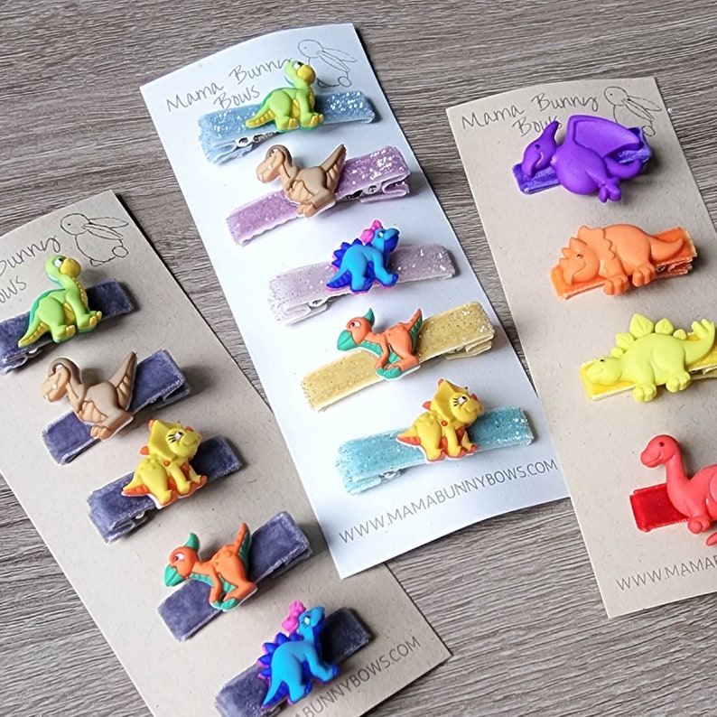 May include: A set of 10 colorful dinosaur hair clips on cards with the text "Mama Bunny Bows" and website address. The clips are made of velvet and glitter and feature different dinosaur designs.