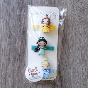 May include: Three princess-themed hair clips with yellow, blue, and green velvet ribbons. The clips feature Belle, Jasmine, and Cinderella. The packaging includes a thank you message for supporting small businesses.