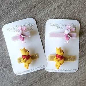 Puede incluir: Dos pinzas para el pelo de Winnie de Pooh. Una pinza presenta un Piglet rosa con una camisa a rayas rosa y blanca y una cinta de purpurina rosa. La otra pinza presenta un Winnie de Pooh amarillo con una camisa roja y una cinta de purpurina dorada. Ambas pinzas est&aacute;n en tarjetas blancas con el texto "Mama Bunny Bows" y una direcci&oacute;n de sitio web.