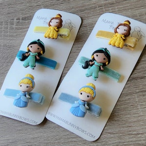 May include: Two sets of princess-themed hair clips. Each set includes a blue, green, and yellow clip with a princess character on each. The clips are on white cards with the text "Mama Bunny Bows" and a website address.