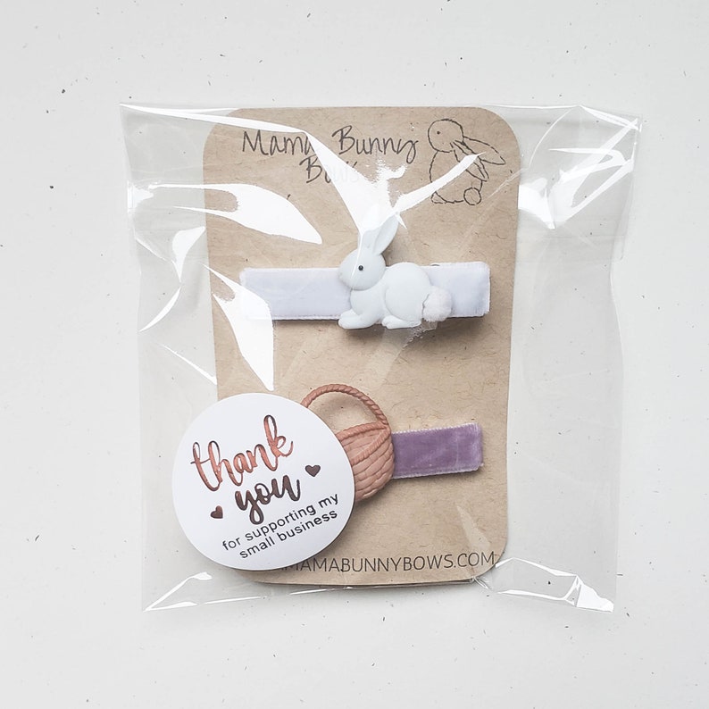 Puede incluir: Dos pinzas para el pelo empaquetadas en una bolsa de pl&aacute;stico transparente. La primera pinza presenta un conejito blanco con una cinta de terciopelo lavanda. La segunda pinza tiene una cesta marr&oacute;n con una cinta de terciopelo morado. Se incluye una pegatina redonda con el texto "gracias por apoyar mi peque&ntilde;o negocio".