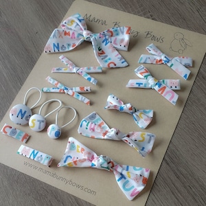 May include: Various hair accessories, including bows, barrettes, and hair ties, are displayed. They are made from white fabric printed with a colourful alphabet. The items are arranged on a brown surface, with the brand name "Mama Bunny Bows" visible.