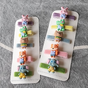 May include: Set of 10 colourful hair clips with various cartoon animal designs, including a crocodile, elephant, fox, and bunny. The clips are attached to velvet ribbons and feature the text "www.mamabunnybows.com".