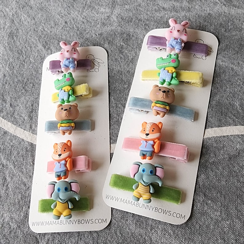May include: Set of 10 colourful hair clips with various cartoon animal designs, including a crocodile, elephant, fox, and bunny. The clips are attached to velvet ribbons and feature the text "www.mamabunnybows.com".