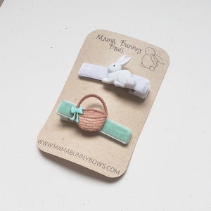 Puede incluir: Dos pinzas para el pelo con un conejito blanco y una cesta marr&oacute;n con un lazo verde. Las pinzas est&aacute;n en una tarjeta marr&oacute;n con el texto "Mama Bunny Bows" y "www.mamabunnybows.com".