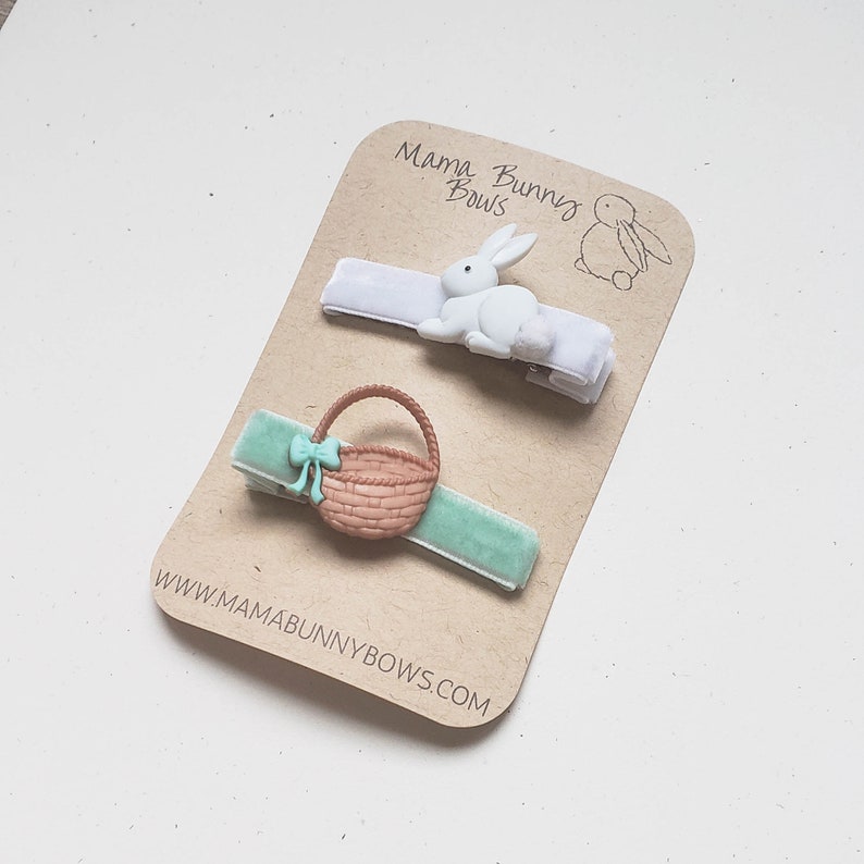 Puede incluir: Dos pinzas para el pelo con un conejito blanco y una cesta marr&oacute;n con un lazo verde. Las pinzas est&aacute;n en una tarjeta marr&oacute;n con el texto "Mama Bunny Bows" y "www.mamabunnybows.com".
