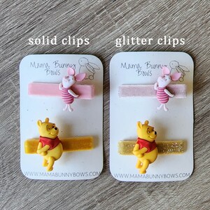 Puede incluir: Dos juegos de pinzas para el pelo con los personajes de Winnie de Pooh y Piglet. El juego de la izquierda tiene pinzas rosas y amarillas s&oacute;lidas, mientras que el juego de la derecha tiene pinzas rosas y amarillas con purpurina. Ambos juegos tienen el texto "Mama Bunny Bows" y una direcci&oacute;n de sitio web.