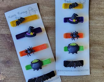 May include: Two sets of Halloween hair clips on white cards. Each set includes a witch's hat, a spider, a cauldron, and a bat. The cards have the text "Mama Bunny Bows" and a website address.