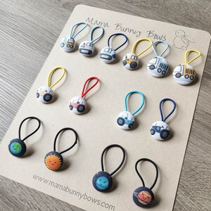 May include: A collection of 12 colorful hair ties with various designs, including construction vehicles and planets. The hair ties are made of elastic and have a fabric covering. The hair ties are arranged on a brown card with the text "Mama Bunny Bows" and a website address.