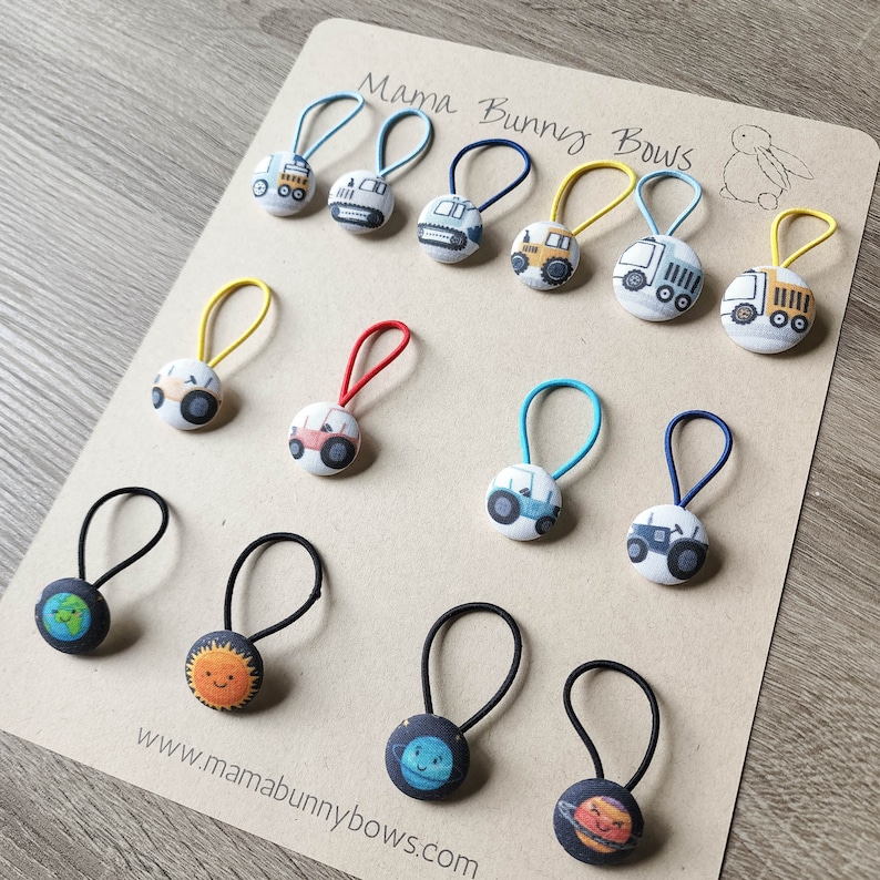 May include: A collection of 12 colorful hair ties with various designs, including construction vehicles and planets. The hair ties are made of elastic and have a fabric covering. The hair ties are arranged on a brown card with the text "Mama Bunny Bows" and a website address.