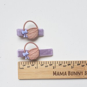 Puede incluir: Dos pinzas para el pelo de terciopelo morado con abalorios de cesta de mimbre marr&oacute;n y lazos de color morado claro. Las pinzas se muestran junto a una regla de madera con marcas en pulgadas.