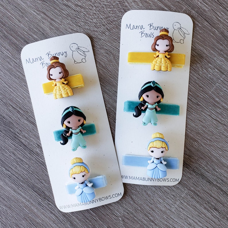 May include: Three sets of princess-themed hair clips with velvet ribbon. The clips feature Belle, Jasmine, and Cinderella. The cards have the text "Mama Bunny Bows" and "www.mamabunnybows.com".