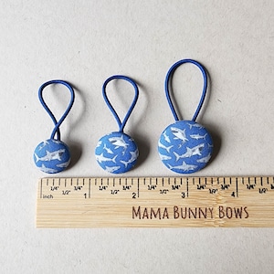 May include: Three blue hair ties with white shark print fabric buttons. The buttons are 1 inch, 1.5 inches, and 2 inches in diameter. The hair ties are on a wooden ruler with the text "MAMA BUNNY BOWS".