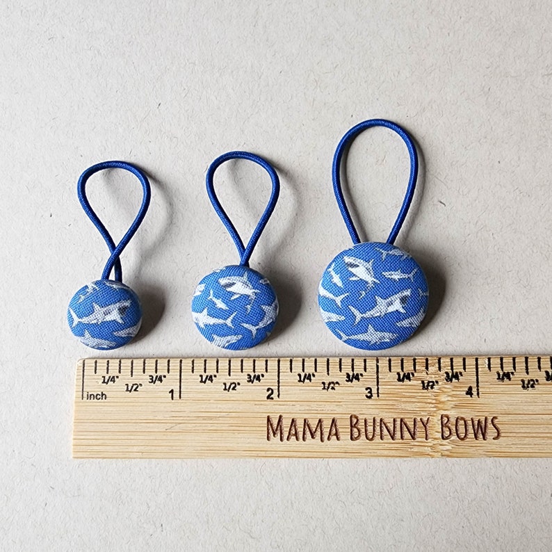 May include: Three blue hair ties with white shark print fabric buttons. The buttons are 1 inch, 1.5 inches, and 2 inches in diameter. The hair ties are on a wooden ruler with the text "MAMA BUNNY BOWS".