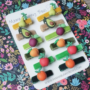 K&ouml;nnte beinhalten: Ein Set mit 12 Haarklammern mit Fruchtmotiven. Jede Klammer zeigt eine andere Frucht, darunter eine Birne, eine Avocado, eine Pflaume, ein Pfirsich, ein Apfel und eine Orange. Die Klammern sind aus Samtband gefertigt und haben eine goldene Metallklammer. Die Klammern sind auf einem wei&szlig;en Hintergrund mit dem Text "Mama Bunny Bows" und "www.mamabunnybows.com" angeordnet.