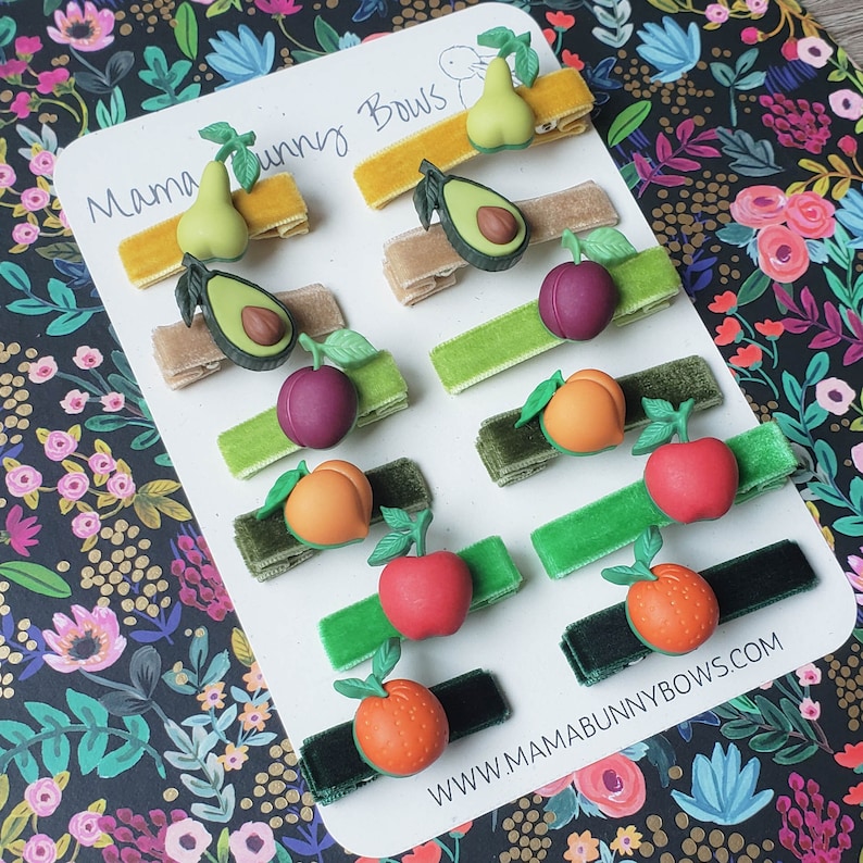 K&ouml;nnte beinhalten: Ein Set mit 12 Haarklammern mit Fruchtmotiven. Jede Klammer zeigt eine andere Frucht, darunter eine Birne, eine Avocado, eine Pflaume, ein Pfirsich, ein Apfel und eine Orange. Die Klammern sind aus Samtband gefertigt und haben eine goldene Metallklammer. Die Klammern sind auf einem wei&szlig;en Hintergrund mit dem Text "Mama Bunny Bows" und "www.mamabunnybows.com" angeordnet.