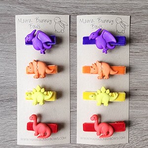 May include: Two cards with the text "Mama Bunny Bows" and a picture of a bunny. Each card has five hair clips with different dinosaur designs. The dinosaurs are purple, orange, yellow, and red. The hair clips are on a brown background.