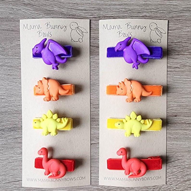 May include: Two cards with the text "Mama Bunny Bows" and a picture of a bunny. Each card has five hair clips with different dinosaur designs. The dinosaurs are purple, orange, yellow, and red. The hair clips are on a brown background.