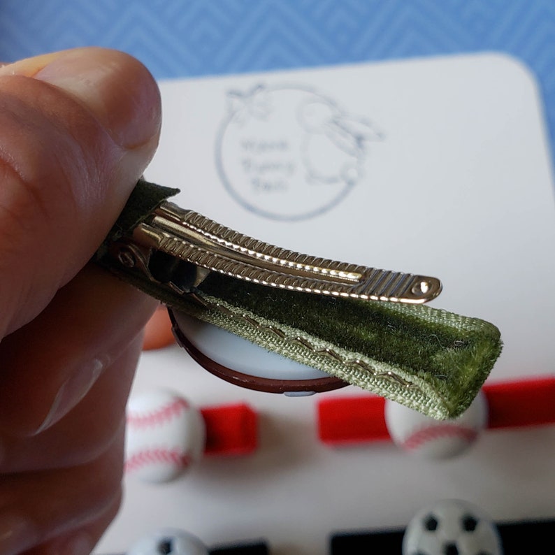 May include: A green velvet hair clip with a gold metal alligator clip. The clip is decorated with a white and brown baseball.