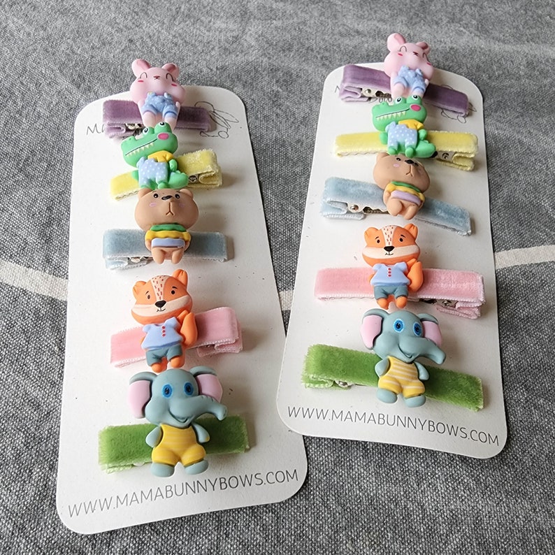 May include: Two sets of colourful hair clips with animal designs. Each set has six clips with different animals, including a bunny, a frog, a bear, a fox, an elephant, and a crocodile. The clips are attached to velvet ribbons in various colours. The bottom of each card reads "www.mamabunnybows.com".