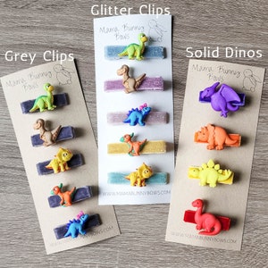 May include: Three cards with different dinosaur hair clips. The first card says "Grey Clips" and has grey velvet clips with green, brown, yellow, and blue dinosaur figures. The second card says "Glitter Clips" and has glitter velvet clips with green, brown, yellow, and blue dinosaur figures. The third card says "Solid Dinos" and has solid velvet clips with purple, orange, yellow, and red dinosaur figures. All cards have the text "Mama Bunny Bows" and a website address.