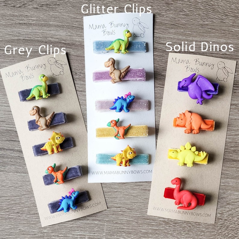 May include: Three cards with different dinosaur hair clips. The first card says "Grey Clips" and has grey velvet clips with green, brown, yellow, and blue dinosaur figures. The second card says "Glitter Clips" and has glitter velvet clips with green, brown, yellow, and blue dinosaur figures. The third card says "Solid Dinos" and has solid velvet clips with purple, orange, yellow, and red dinosaur figures. All cards have the text "Mama Bunny Bows" and a website address.