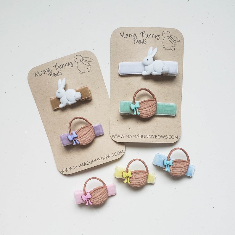 Puede incluir: Conjunto de seis pinzas para el pelo con tem&aacute;tica de Pascua con un conejito blanco y una cesta marr&oacute;n con un lazo. Las pinzas est&aacute;n en una tarjeta marr&oacute;n con el texto "Mama Bunny Bows" y la direcci&oacute;n del sitio web "www.mamabunnybows.com".