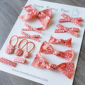 May include: A set of coral pink hair bows with a white cross pattern. The set includes a large bow, smaller bows, and hair ties with matching buttons. The bows are made of fabric and have a knotted centre. The text "Mama Bunny Bows" is written on the white background.