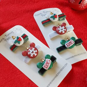 K&ouml;nnte beinhalten: Satz mit f&uuml;nf Weihnachts-Haarklammern mit roten und gr&uuml;nen Ornamenten mit Schneeflocken- und "Noel"-Designs. Die Klammern befinden sich auf wei&szlig;en Karten mit dem Text "Mama Bunny Bows" und einer Website-Adresse.
