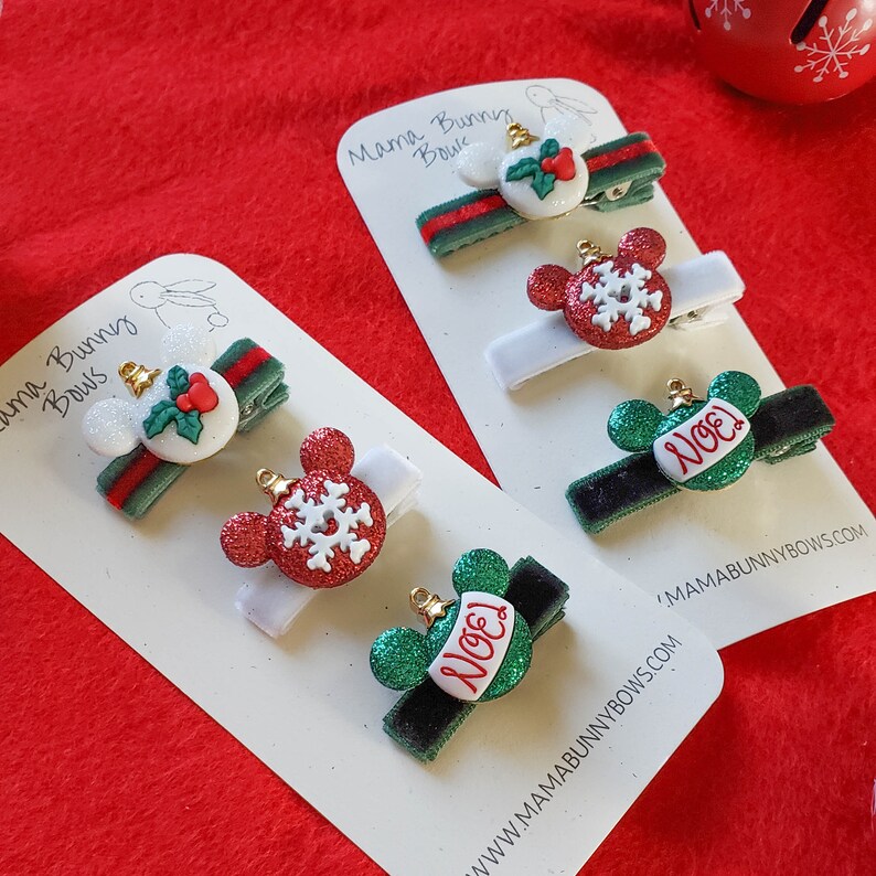 K&ouml;nnte beinhalten: Satz mit f&uuml;nf Weihnachts-Haarklammern mit roten und gr&uuml;nen Ornamenten mit Schneeflocken- und "Noel"-Designs. Die Klammern befinden sich auf wei&szlig;en Karten mit dem Text "Mama Bunny Bows" und einer Website-Adresse.