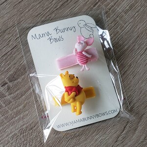 Puede incluir: Dos pinzas para el pelo con personajes de Winnie the Pooh. Una pinza presenta a Piglet con un lazo rosa y la otra pinza presenta a Winnie the Pooh con un lazo rojo. Las pinzas est&aacute;n empaquetadas en una tarjeta blanca con el texto "Mama Bunny Bows" y la direcci&oacute;n del sitio web "www.mamabunnybows.com".