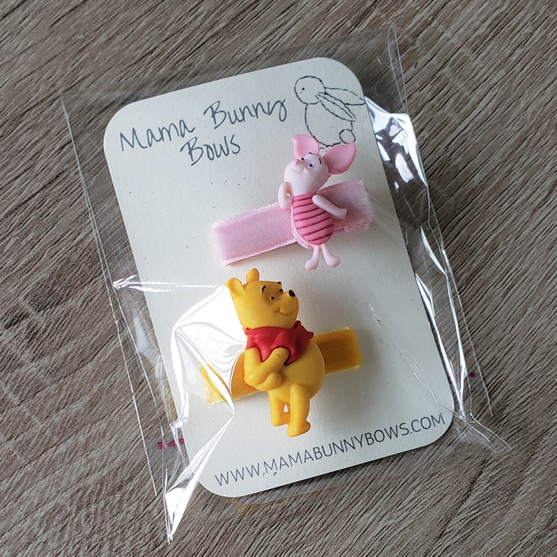 Puede incluir: Dos pinzas para el pelo con personajes de Winnie the Pooh. Una pinza presenta a Piglet con un lazo rosa y la otra pinza presenta a Winnie the Pooh con un lazo rojo. Las pinzas est&aacute;n empaquetadas en una tarjeta blanca con el texto "Mama Bunny Bows" y la direcci&oacute;n del sitio web "www.mamabunnybows.com".