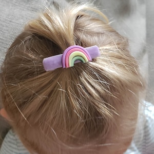 May include: A purple hair clip with a rainbow design. The rainbow is made of five colors: pink, yellow, green, light blue, and dark blue.