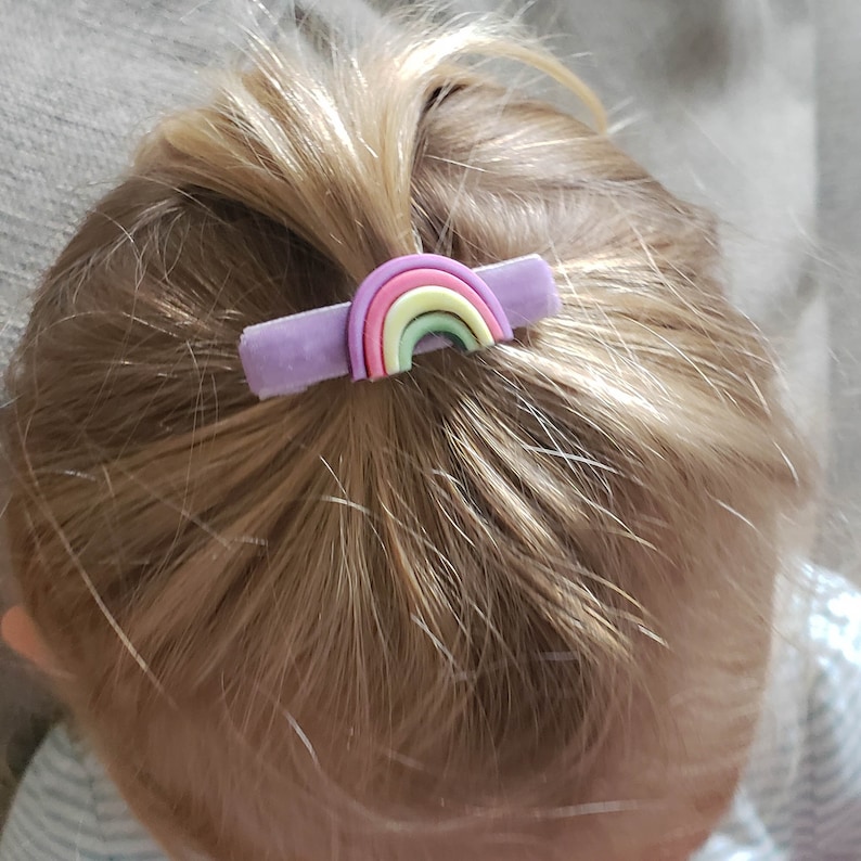 May include: A purple hair clip with a rainbow design. The rainbow is made of five colors: pink, yellow, green, light blue, and dark blue.