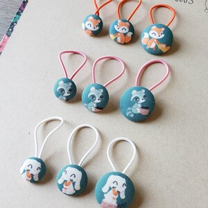 May include: A set of nine hair scrunchies with different animal designs. The scrunchies are made of fabric and have a button-like design. The animals include foxes, raccoons, and bunnies. The scrunchies are in various colors, including pink, white, and orange.