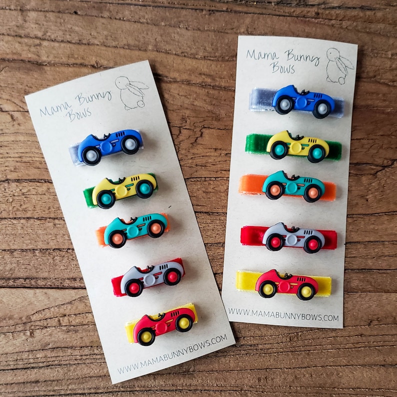 May include: Set of six colorful car hair clips with velvet ribbon. The cars are blue, yellow, green, gray, red, and red. The ribbon colors are gray, green, orange, red, yellow, and red. The clips are displayed on a card with the text "Mama Bunny Bows" and "www.mamabunnybows.com".