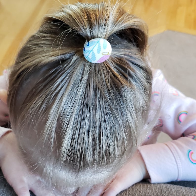May include: A young girl with blonde hair wearing a pink shirt with rainbow designs has her hair pulled back in a ponytail with a blue and white patterned fabric hair tie.