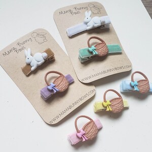 Puede incluir: Conjunto de seis pinzas para el pelo con dise&ntilde;o de conejo y cesta. Las pinzas est&aacute;n en tarjetas marrones con el texto "Mama Bunny Bows" y la direcci&oacute;n del sitio web "www.mamabunnybows.com".