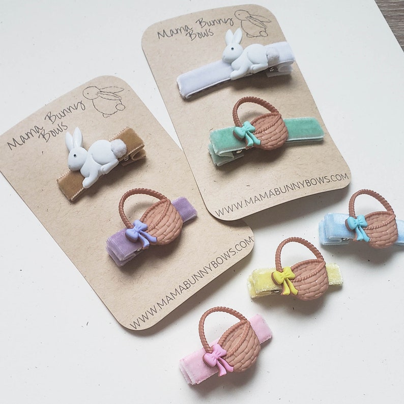 Puede incluir: Conjunto de seis pinzas para el pelo con dise&ntilde;o de conejo y cesta. Las pinzas est&aacute;n en tarjetas marrones con el texto "Mama Bunny Bows" y la direcci&oacute;n del sitio web "www.mamabunnybows.com".